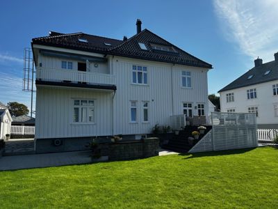 Fotografi av en hvit, toetasjes villa sett fra bakgården. Bildet viser fasaden, taket, trappen opp til inngangen, balkongen og omgivelsene.