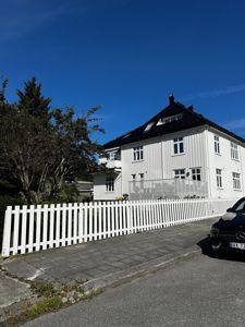 Fotografi av en hvit, trepanelt bygning med svart tak sett fra gaten. Bildet viser fasaden, en hvit stueplass, en bil og en gate.