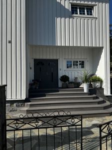 Fotografi av en hvit, vertikal paneltett fasade med en mørk dobbeltdør, trapper og planter.