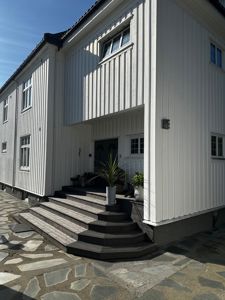 Fotografi av en hvit, trepanelert bygning sett fra siden, med trapper opp til inngangsdøren og en steinlagt fortau.