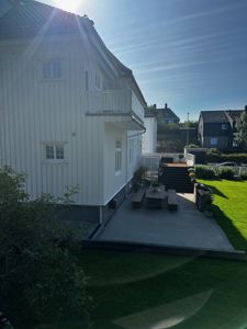 Fotografi av en hvit, trepanelert bolig sett fra bakgården. Bildet viser fasaden, et balkong, en terrasse med møbler og en trapp opp til en hage. Det er også synlig naboer og sollys.