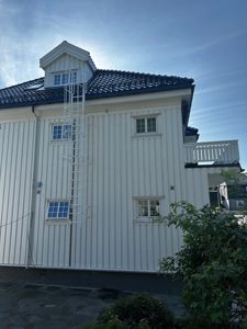 Fotografi av fasaden på en hvit, trepanelert bygning med etasjer, vinduer, en takterrasse og en hvit branntrappe.