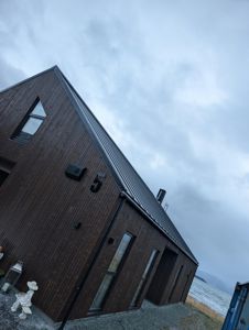 Fotografi av en ferdigbygd eller under oppføring stående bygning med mørk trefasade og metalltak, sett fra et lavt vinkel mot en skyet himmel.