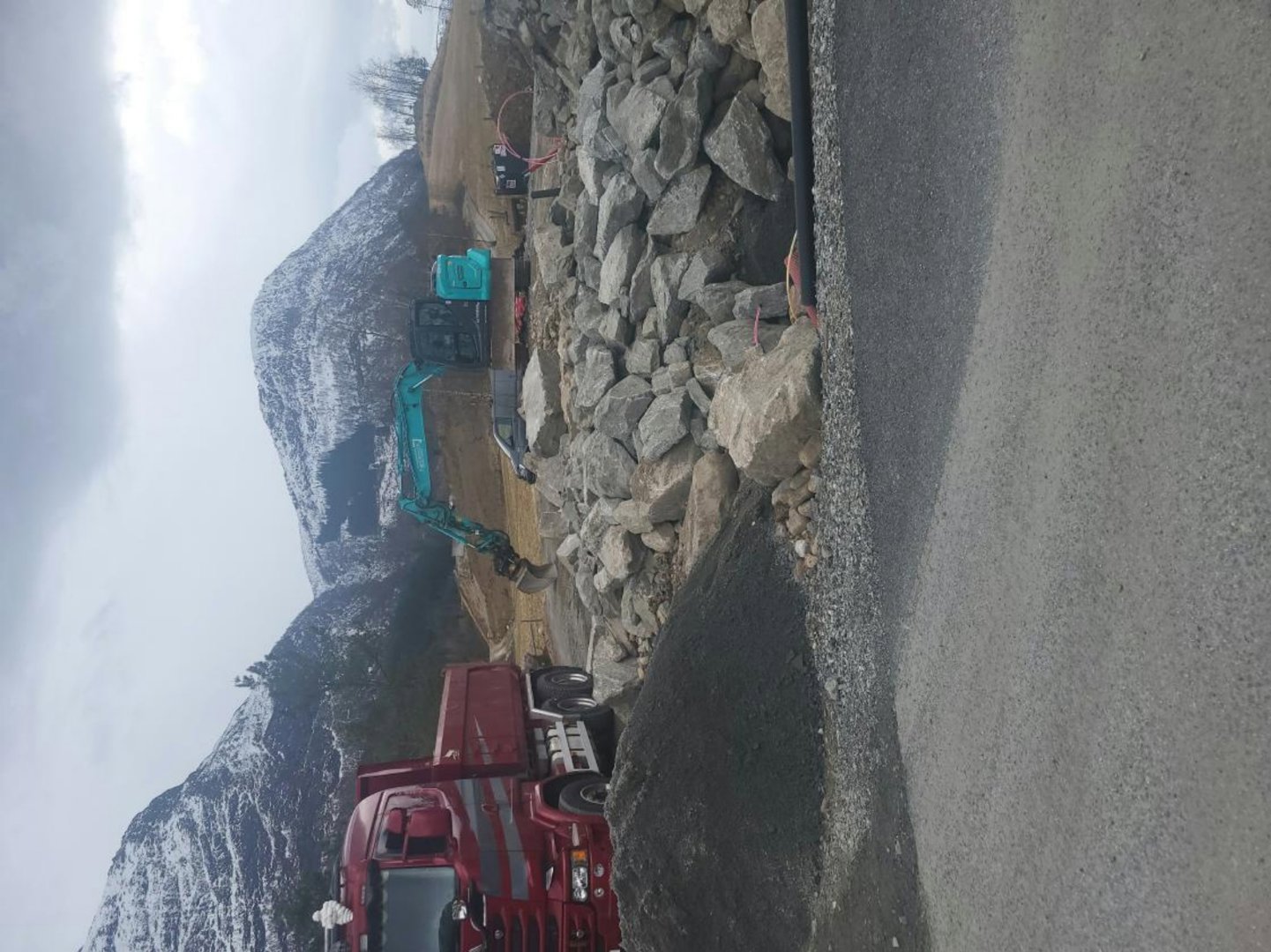 Fotografi av en byggeplass med tungt utstyr (graver, lastebil) og steinmasser i et fjellområde.