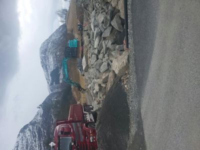 Fotografi av en byggeplass med tungt utstyr (graver, lastebil) og steinmasser i et fjellområde.