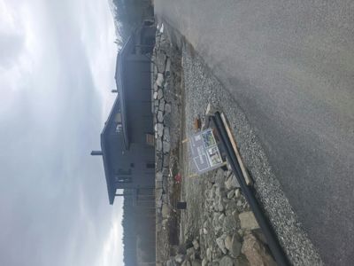 Fotografi av en mørk bygning under oppføring eller ferdigstillelse, sett fra siden med en steinmur og en vei i forgrunnen.