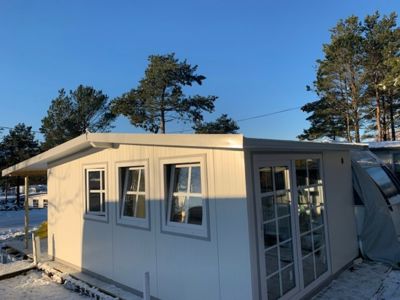 Fotografi av en ferdig eller nesten ferdig bygning (hvit panelvegg) sett fra siden, med snø på bakken og trær i bakgrunnen.