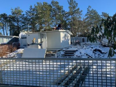 Fotografi av en hvit container som står på et snødekket område, med arbeidere på taket og en gitterhekk i forgrunnen.