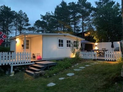 Fotografi av en hvit, moderne hytte eller containerbolig sett fra siden, med belyst veranda, hage og omgivende skog.