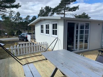 Fotografi av en hvit containerhytte med en terrasse, hvit hekk og en bil i bakgrunnen.