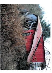Fotografi av en gammel, rød fasettbygning (tømmerhytte) som står på en skråning og er omgitt av busker og trær.