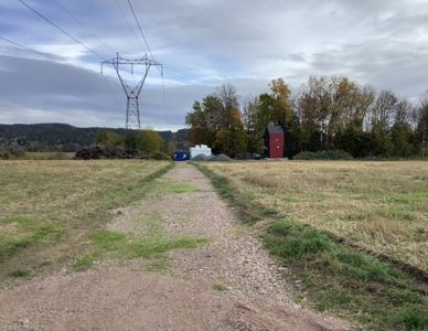 Fotografi av en tomt med en liten rød bygning, en grusvei, og en høyspenningspære i bakgrunnen.