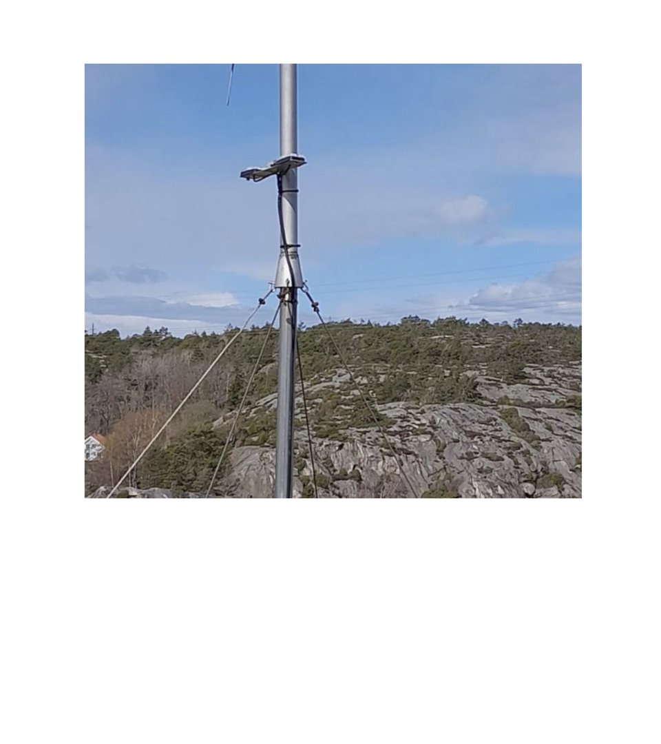 Fotografi av en metallmast eller antenne på en steinete bakke med vegetasjon og himmel i bakgrunnen.