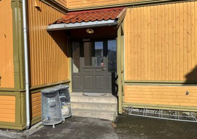 Fotografi av en bygning med gule fasader, en grå inngangsdør, trapper og en liten takoverheng.