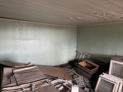 Fotografi av et rom inne i en bygning, sannsynligvis et kjellerrom eller et lager, som er fylt med diverse gjenstander og ser ut til å være i dårlig stand.