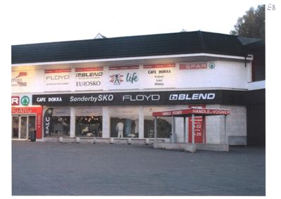 Fotografi av en eksisterende bygning (handelsbygg) med synlige butikkutsikter og skilt.