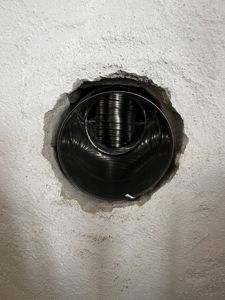 Fotografi av en rund ventilasjonsåpning i en hvit, kornete vegg. Bildet viser en svart, spiralformet ventilasjonsrør som sitter i et hull i veggen.