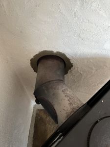 Fotografi av en metallrørledning (kanskje peisrør eller ventilasjon) som går gjennom et hull i en hvitmalt betong- eller gipsvegg.