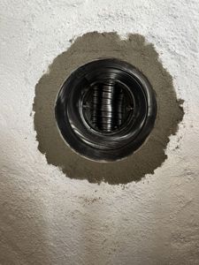 Fotografi av en installert ventilasjonsrør i en hvit, kornet vegg. Røret er omgitt av grå mørtel.