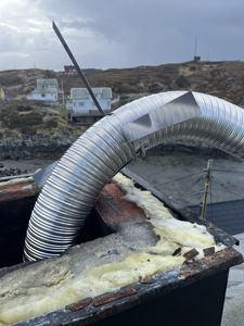 Fotografi av en fleksibel metallrørledning (trollrør) som er montert gjennom et tak med isolasjon, med utsikt over et kystlandskap i bakgrunnen.
