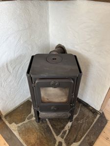 Fotografi av en svart vedovn plassert i en hjørne med hvite, klossede vegger og en steinbelagt gulvplate.