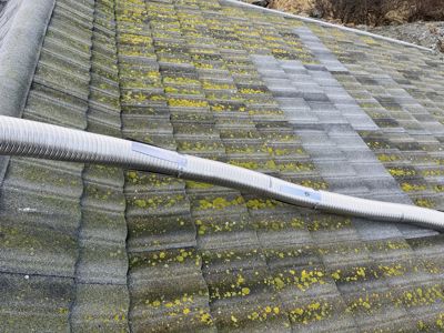 Fotografi av et tak med grå takfliser og gulgrønn mos, med en fleksibel metallrørledning (muligens ventilasjon) som ligger på taket.