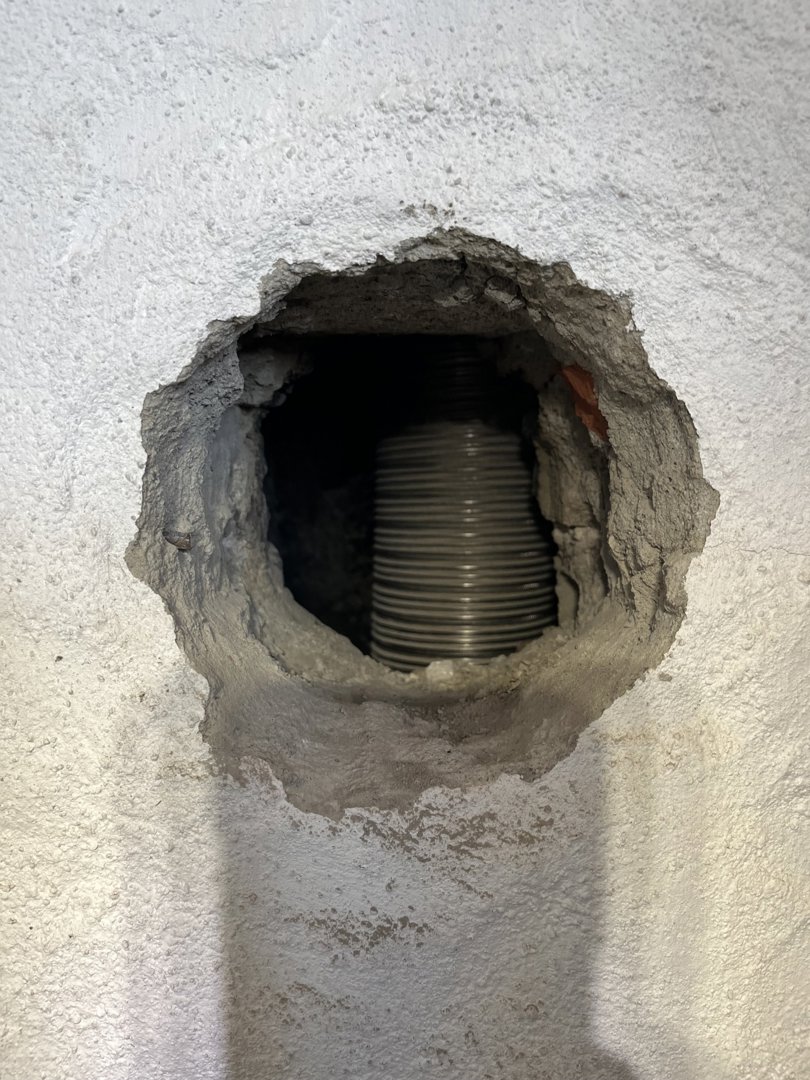 Fotografi av en hull i en hvitputtet vegg som viser en grå, ribbet rørledning (sannsynligvis ventilasjonsrør) som går gjennom veggen.