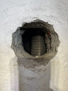 Fotografi av en hull i en hvitputtet vegg som viser en grå, ribbet rørledning (sannsynligvis ventilasjonsrør) som går gjennom veggen.