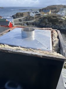 Fotografi av en metallisk taklukking (t.d. for skorstein eller ventilasjon) med isolasjon, sett fra et tak med hav og hus i bakgrunnen.