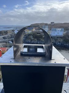 Fotografi av en metallisk ventilasjons- eller røykledning på taket, med utsikt over et kystnær landskap i bakgrunnen.