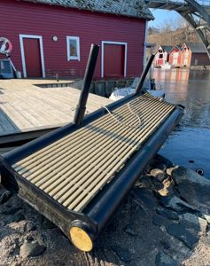 Fotografi av en rød bygning ved vannet med en brygge og et båtbygg i forgrunnen.