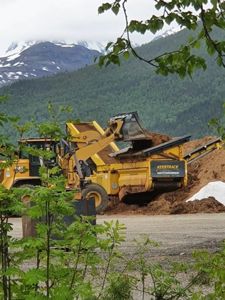 Fotografi av en byggeplass med grøftemaskiner (KeeStrack) i arbeid, med fjell og skog i bakgrunnen.