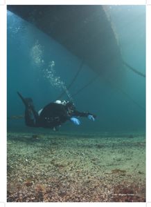 Fotografi av en dykker under vann, med en stor skipsskrog i bakgrunnen.