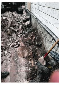 Fotografi av en byggeplass eller utbedringsarbeid ved en bygning, som viser graving, stein og en spade.