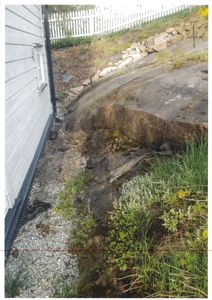 Fotografi av bygningens sidevegg, en steil bakke med grus og vegetasjon, samt en hvit hekk i bakgrunnen.