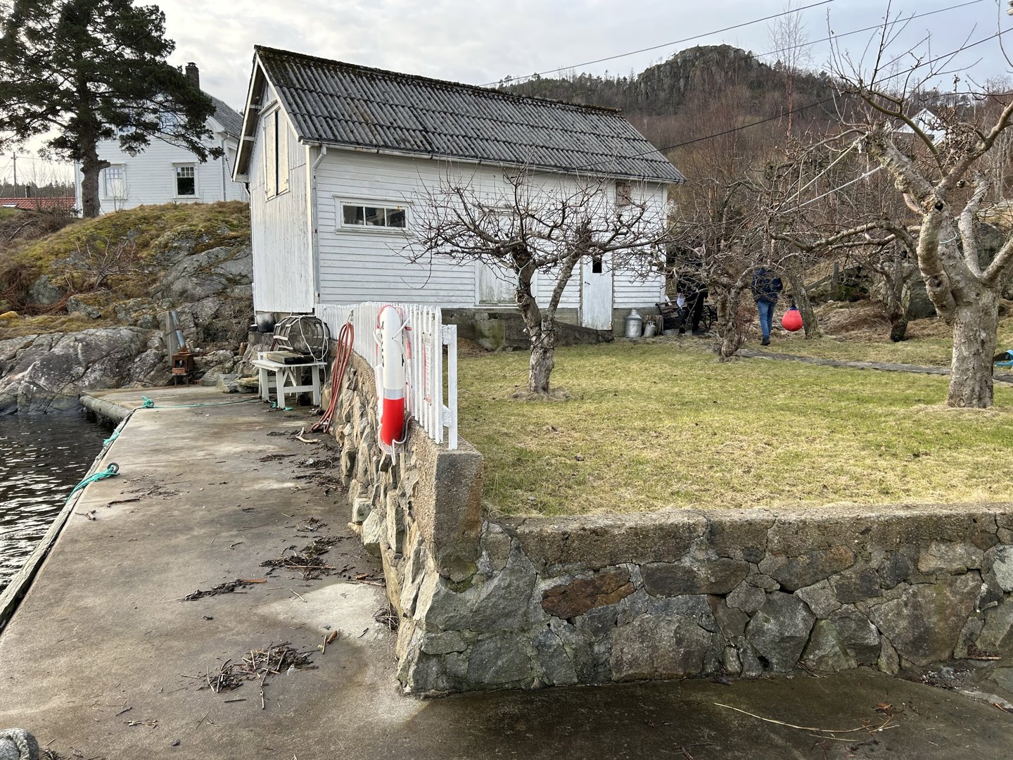 Fotografi av en hvit, gammel bygning ved kysten med en steinmur og en kai i forgrunnen.