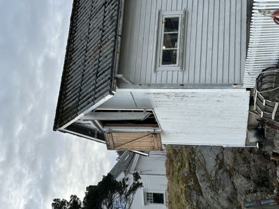 Fotografi av en hvit, trepanelert bygning med et tak som ser ut til å være i dårlig stand (moss/mo). Bildet viser fasaden og hjørnet av huset.
