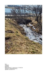 Fotografi av et utsnitt av et terreng med en bekke, snøflekker og trær, med en vei og vann i bakgrunnen.