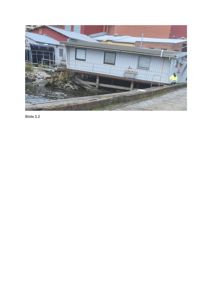 Fotografi av en hvit, langhuslik bygning på en kai ved vannet, med en person i gule vesten i bakgrunnen.