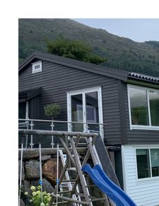 Fotografi av en mørk, moderne hytte med hvite vinduer, sett fra bakken mot en bakke.