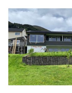 Fotografi av en bolig under ombygging eller nybygging, med synlig fasade, vinduer og en steinmur i forgrunnen.