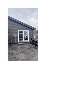 Fotografi av en grå hytte med hvit dør og vinduer, sett fra bakgård med steinbelagt terrasse og potteplanter.