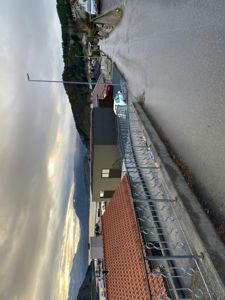 Fotografi av en eksisterende bygning (en grå container-lignende struktur) sett fra gaten, med omgivende landskap, vei og andre hus i bakgrunnen.