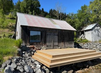 Fotografi av en eksisterende eller ferdigstilt trehytte med et nytt, moderne treterrasse foran. Bildet viser bygningen i sin omgivelse med steinmur og skog.
