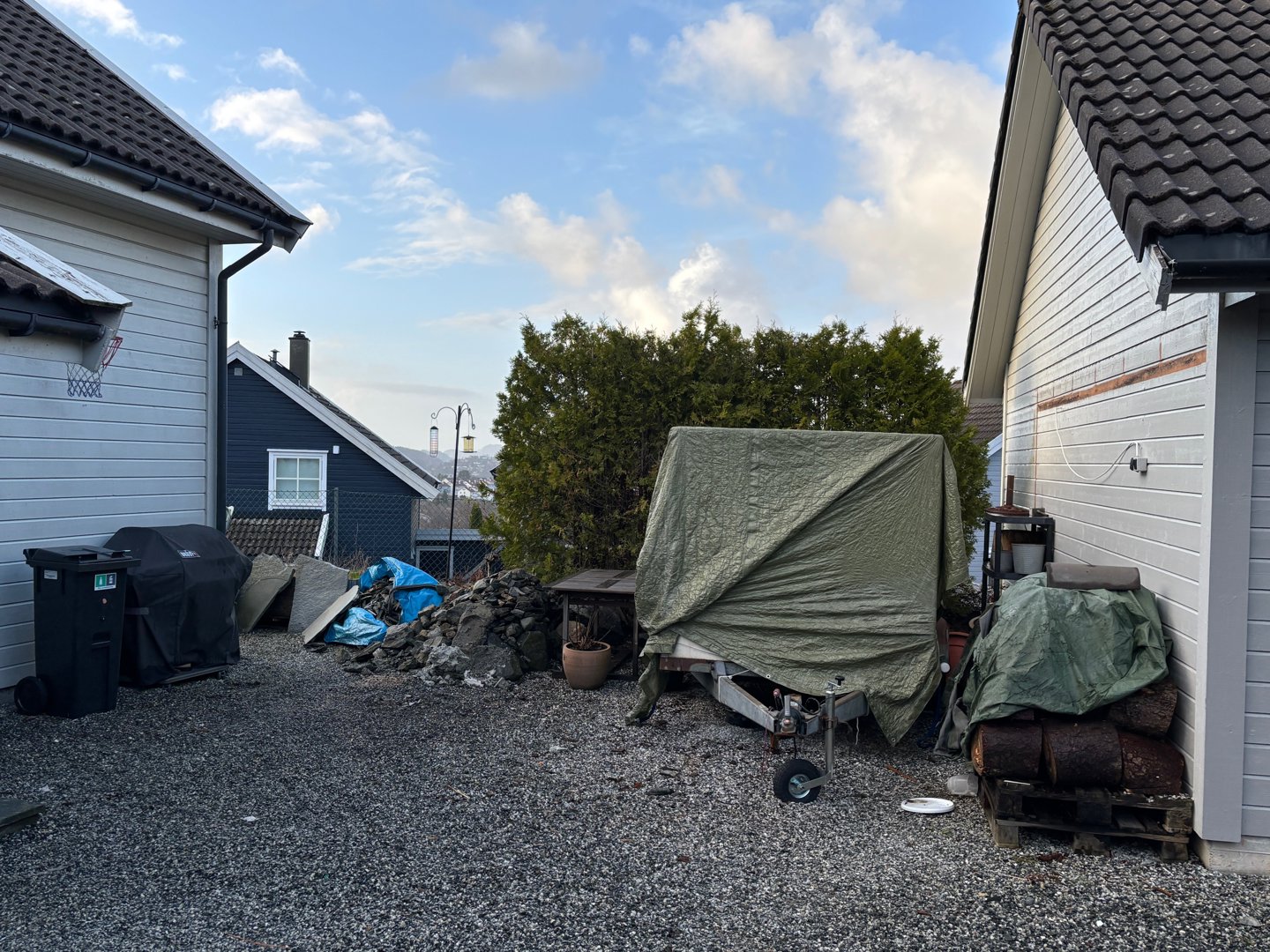 Fotografi av en bakgård eller hageområde mellom to hus, med diverse gjenstander som oppbevares under telt.