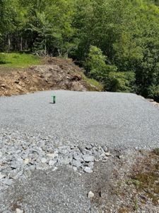 Fotografi av en utgravet og gruslagt tomt med et grønt markørstang i midten, omgitt av skog.