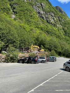 Fotografi av en byggeplass eller nedrivningsarbeid ved en vei, med en gulelastbil og en delvis nedrevet struktur i bakgrunnen mot en fjellside.