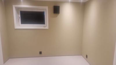 Fotografi av et tomt, innredningsfritt rom med en vindu og en veggmontert høyttaler.