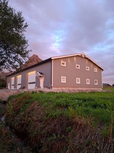 Fotografi av en ferdigbygd eller nesten ferdigbygd bygning (gjerne en uteskure eller garasje) med grå fasade, hvite dører og vinduer, sett fra et lavt vinkel med en bakke i forgrunnen og en fjelltopp i bakgrunnen.