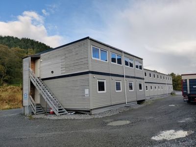 Fotografi av en rekke av toetasjers modulbygg (kanskje containerboliger) på en grusplass med skog i bakgrunnen.